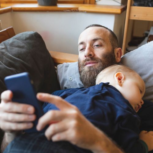 man looking at phone whilst baby sleeps on him